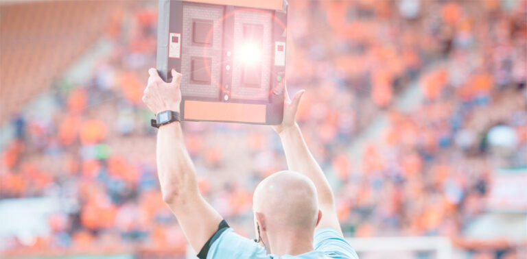 Árbitro asistente levantando un tablero electrónico para anunciar el tiempo de descuento durante un partido de fútbol.